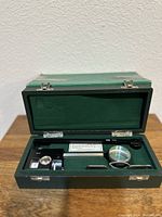 Opened green hinged presentation case with compensating polar planimeter inside, showing black and metal precision parts including magnifier and adjustable arm