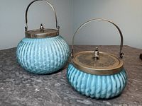 Pair of two blue satin glass biscuit barrels with ribbed surface and silver plated lids and swing handles. Larger barrel is taller with a chip on the glass.