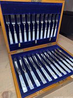 Open wooden presentation case with blue velvet lining showing 12 forks in the lid and 12 knives in the base