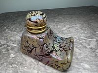 Front perspective of iridescent glass inkwell showing boat-like shape, black spatter decoration, metal screw-on lid closed