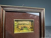 Framed miniature artwork showing a landscape with a stone bridge, trees, and a small building. The artwork includes gold foil accents. The frame is wood with a gold inner trim.