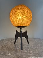 Front view of mid century table lamp lit, showing glowing textured amber plastic shade and tripod teak wood base with pull chain switch.