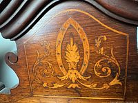 Close-up of top pediment showing intricate marquetry inlay in a shield and scroll motif on walnut-stained wood.
