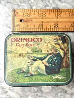 Top view of rectangular tin showing the printed image of a man in colonial clothing smoking a pipe, the words 'Orinoco Cut Fine' printed in red on the lid along with a wooden ruler above for size reference.
