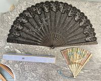 Two antique Victorian fans laid open; one is a black lace mourning fan with hand-carved wooden sticks, the other a smaller hand-painted floral fan with tassel attached.