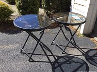 Two round glass-topped foldable patio tables shown outdoors on pavement with sunlight casting shadows. Metal frames are black with slight scratches and there is garage dust visible.