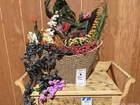 Full basket view showing various faux flowers including tropical broad green leaves and small colorful flower clusters in reds, purples, and yellows.