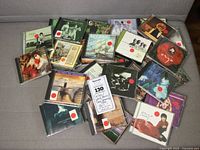 Photo showing 20 assorted CDs in jewel cases scattered on gray fabric surface. Red circular price stickers on each case.
