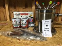 Stainless steel fondue pot with six colorful fondue forks, cans of Sterno cooking fuel, and extra forks in plastic bag on wooden surface.