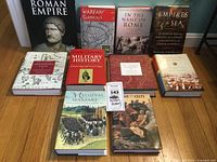 Full view of 10 military history books arranged on the floor with a Roman bust in the background. Various titles on classical, medieval, and empire military history.