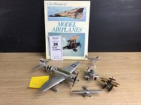Photo showing the five model airplanes arranged on a wooden table with the book 'Color Treasury of Model Airplanes' behind them, showing various styles of planes including WWII fighter and vintage models.