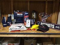 Shelf display of various New England Patriots fan items including apparel like hat and apron, several beer cozies, Ziploc bags, a commemorative football, a small framed display, and decorative street sign.