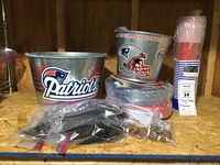 Photo of two galvanized metal pails with New England Patriots logos, stacked plates, and bags of silver plastic utensils all arranged on a wooden shelf.
