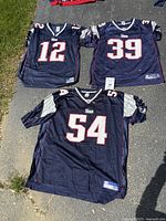 Photo showing three New England Patriots jerseys laid out on the ground in sunlight. Visible player numbers 12 (Brady), 39 (Woodhead), and 54 (Bruschi) with Patriots logos on the shoulders.