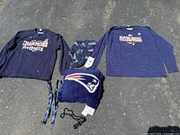 Two 2XL Patriots jerseys, plush pillow, scarf, lanyards and beads displayed on pavement.