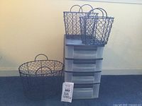 Photo showing three metal wire nesting baskets stacked and placed on top of the four-drawer plastic storage unit against a plain wall, with measurement info visible on small tag in front.