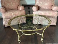 View of the brass and glass coffee table with scalloped-edge top and curved legs with scroll feet, set between two upholstered pink armchairs.