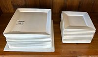Stack of white square porcelain plates showing small, medium, and large sizes from top and side angles, arranged on a wooden shelf.