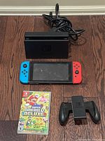 Full lot on hardwood floor: Switch dock with attached cables at rear, console with Joy-Cons, game case and Joy-Con grip in front