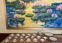 Full view of cups and saucers including multiple plates set on table with large floral painting in background.