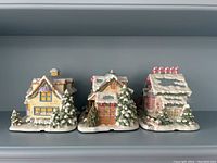 Front view of three Precious Moments ceramic village houses with snowy roofs and holiday decorations.