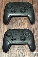 Pair of black Nintendo Switch Pro Controllers shown on wooden floor, front view showing buttons and sticks.