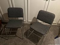 Two grey upholstered office chairs with stainless steel sled frames photographed side by side on carpet in front of white cabinets.