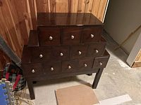Full view of the wooden apothecary CD holder cabinet with 12 drawers arranged in stepped tiers, placed in a room with wood-paneled walls and concrete floor.