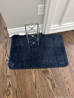Navy blue bath mat placed on hardwood floor with metal toilet paper holder on top.