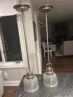 Two pewter-colored pendant lights standing upright with clear textured glass shades and visible wiring, shown near a window and living room background.