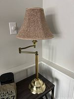 Front view of a tall brass swivel table lamp with beige fabric bell shade on dark wood table against white wall outlet, unlit.