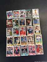 Top-down view of 25 assorted baseball cards laid out in five rows of five on a dark surface. Cards show players in various poses, colorful borders, and different team uniforms.