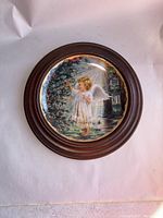 Photo of the framed decorative plate showing the entire piece with the angel illustration, dark brown wooden frame, and white background.