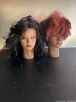 Two mannequin heads side by side; one with black hair and green eyes, the other with reddish-brown hair and brown eyes, both showing facial details and hair texture.