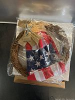 Wreath laying flat with plastic packaging covering it, showing intertwined twig base, American flag fabric, burlap bow, and metal star in center top.