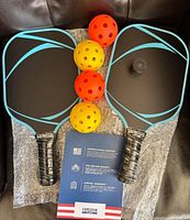 Two black pickleball paddles with blue design and textured grips, four orange and yellow perforated balls, warranty card showing American small business and lifetime warranty text