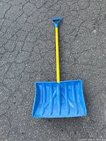 Full view of snow shovel leaning on asphalt showing blue plastic scoop, yellow shaft, and blue handle.