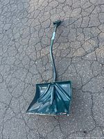 Full view of the green plastic snow shovel lying on pavement showing curved handle and scoop with metal edge.