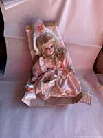 Full doll shot seated in cardboard support with visible pink satin dress and bonnet.