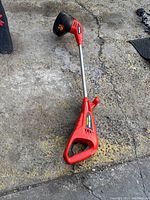 Full view of red cordless weed trimmer with ergonomic handle and shaft on concrete surface.