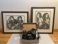 Framed art prints placed against wall with box of cameras, binoculars, and Garmin device in front