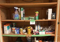 Wooden cupboard shelf with assorted cleaning and home repair supplies including spray cleaner, adhesive tubs, tapes, gloves, bleach cleaner.