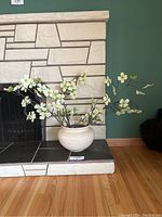 Showcases artificial dogwood flowers in textured round vase on fireplace hearth with stone background.