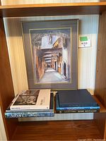 Framed print of an arched corridor and stacked coffee table books below, showing entire display in wood shelf.