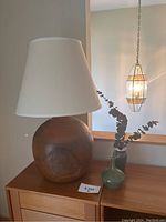 Pottery table lamp with round brown base and white lampshade, two small pottery vases on wooden cabinet near window with hanging lantern outside.