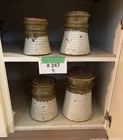 Set of four pottery storage jars arranged inside cupboard shelf. Jars have two-tone glaze and cork lids.