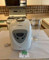 Front view of white Sunbeam bread machine sitting on kitchen counter next to a clear glass for scale. Oven and microwave in background.