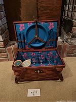 Open top view showing four blue plastic plates secured to the lid, blue-handled cutlery, four clear glasses, a candle, and a coaster inside the basket with floral lining.