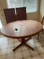 Round teak table top halves joined, green lot tag on surface, pedestal base visible