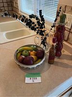 Ceramic bowl with wooden fruit, tall bottle with red objects, smaller bottle with dried beans, and dark leafy branches in vase on kitchen counter.